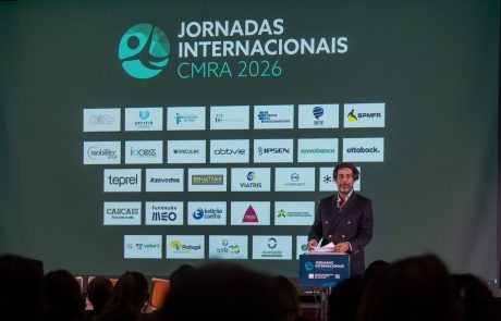Orador intervém ao púlpito perante o ecrã das Jornadas Internacionais de Reabilitação CMRA 2026, com os logótipos das entidades parceiras e patrocinadoras.