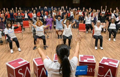 Sessão de Zumba Gold no auditório do Centro de Medicina de Reabilitação de Alcoitão, com dezenas de participantes, alguns em cadeiras de rodas, a acompanhar os movimentos da instrutora com os braços levantados.